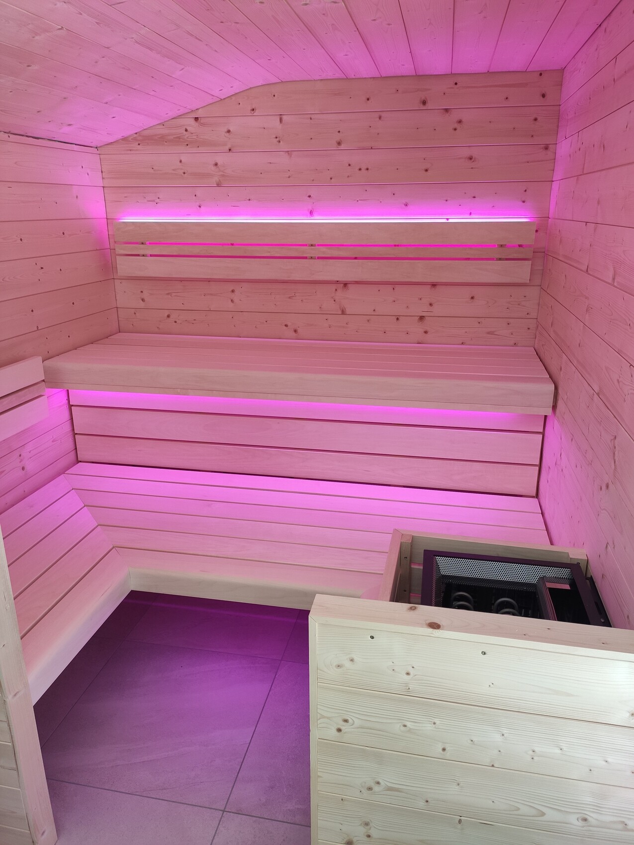Wood sauna with L-shaped benches, heater in corner, pink LED light.