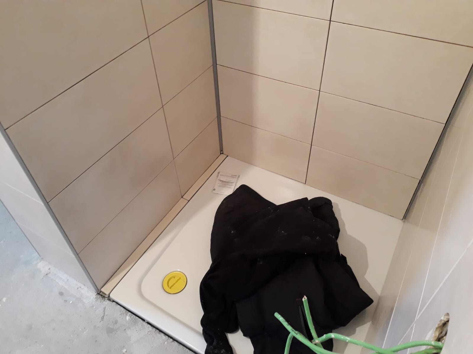 Shower under construction: beige tiles, white shower base, yellow drain cover, black jacket.
