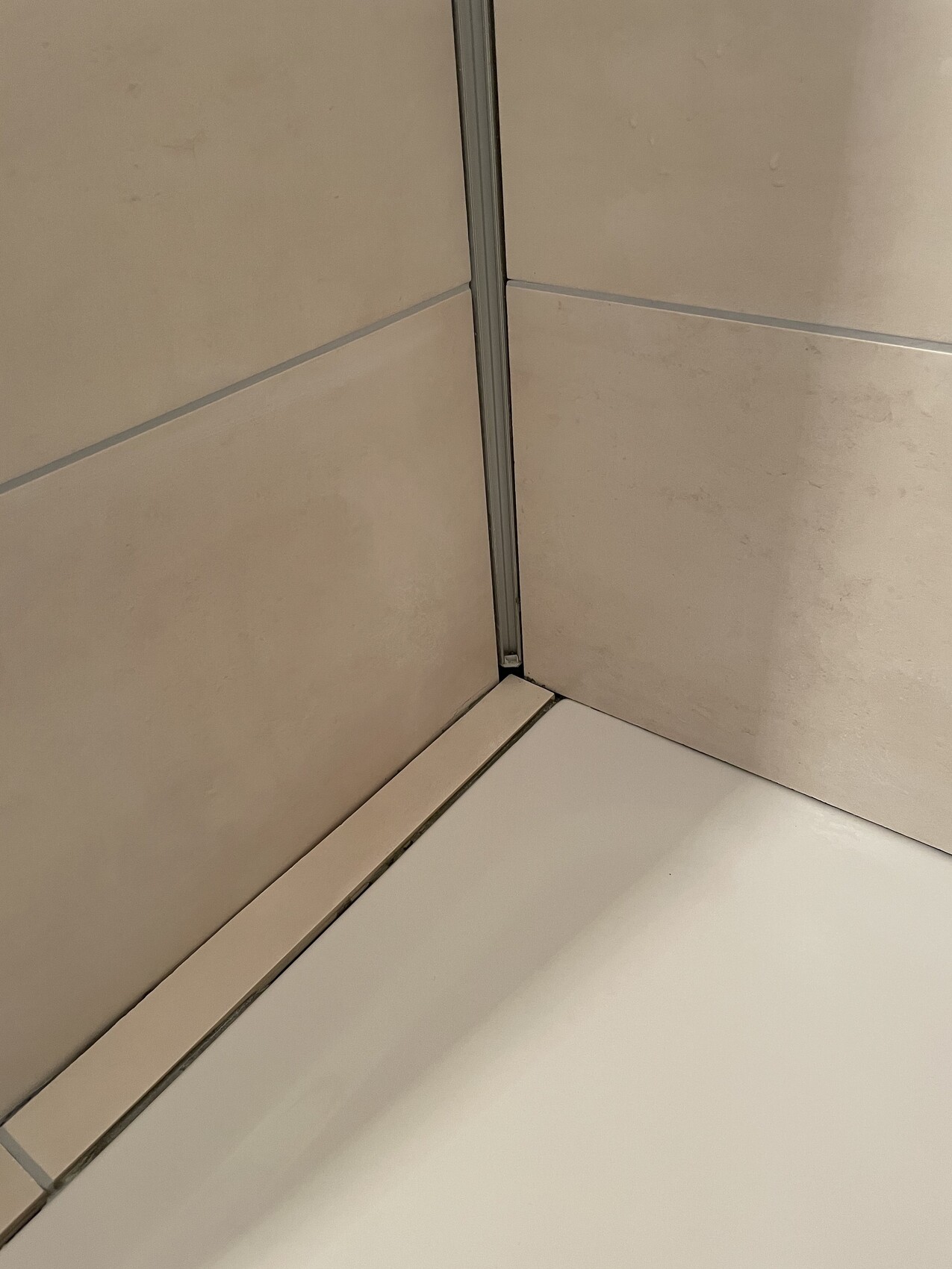 Corner shower with beige tiles, metal profile in the corner, and white shower floor.