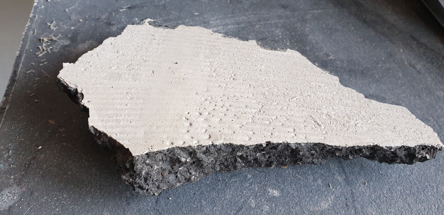 Broken piece of concrete with light, smooth top and dark, porous underside on work surface.