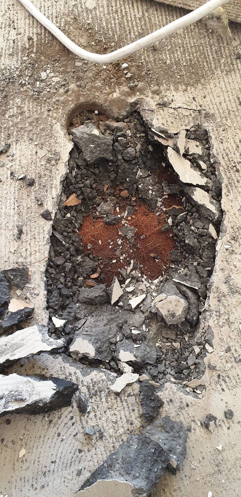 Open hole in concrete floor with broken concrete and rusty reinforcement steel, debris, cables above.