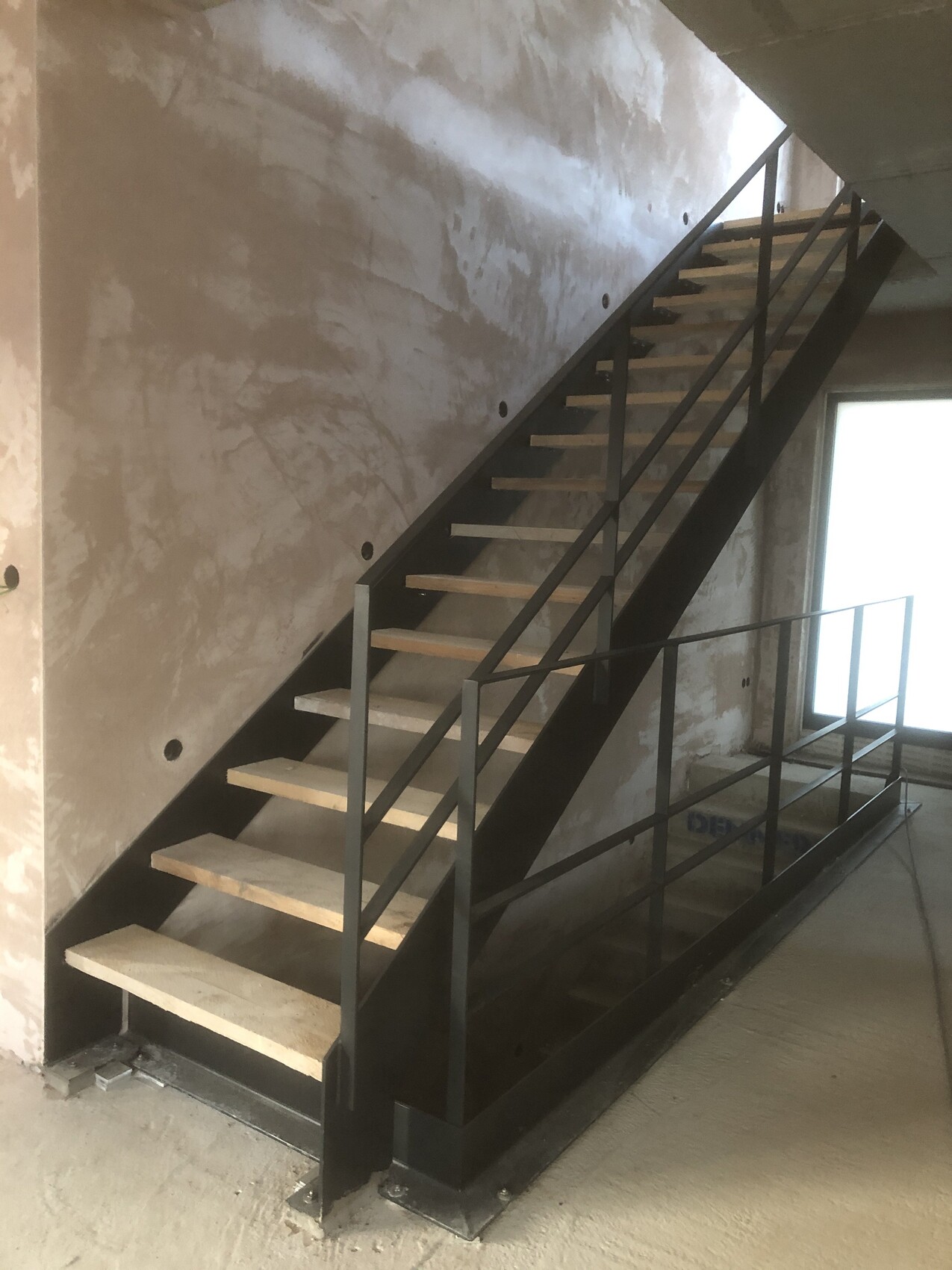 Metal staircase with open wooden steps in unfinished interior, raw masonry and window on the right.