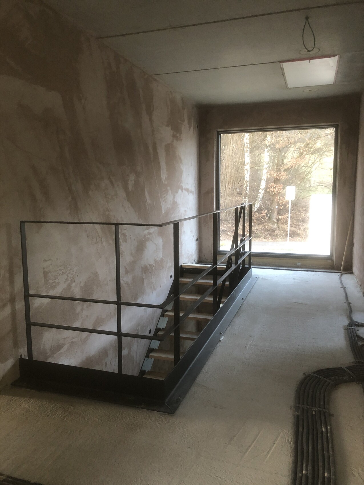 Unfinished interior with steel staircase, rough walls, cables on the floor and large window to the outside.