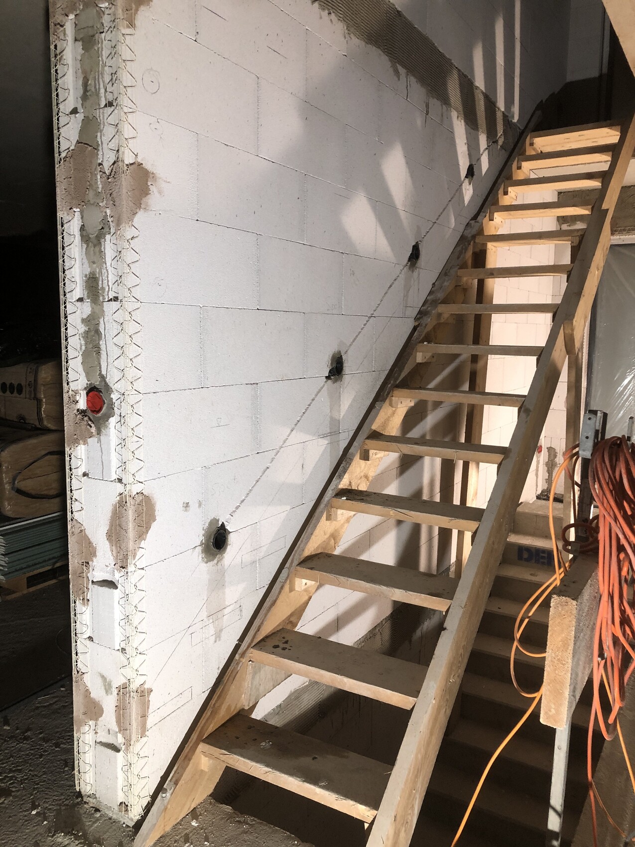 Wooden staircase leaning diagonally against white block wall in shell construction, orange extension cable bottom right.