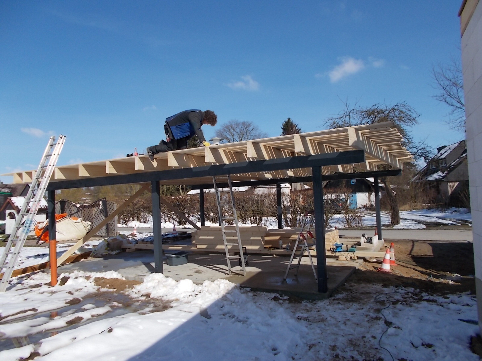 Bauarbeiter montiert Holzdach an Stahlrahmen eines Carports im Schnee, Leiter daneben.