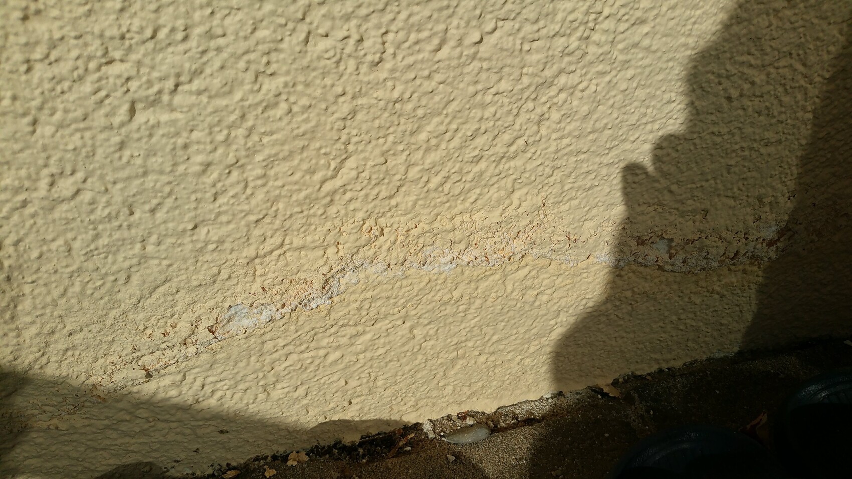 Beige plastered wall with a lower crack line; shadow of a person on the right; ground with gravel.