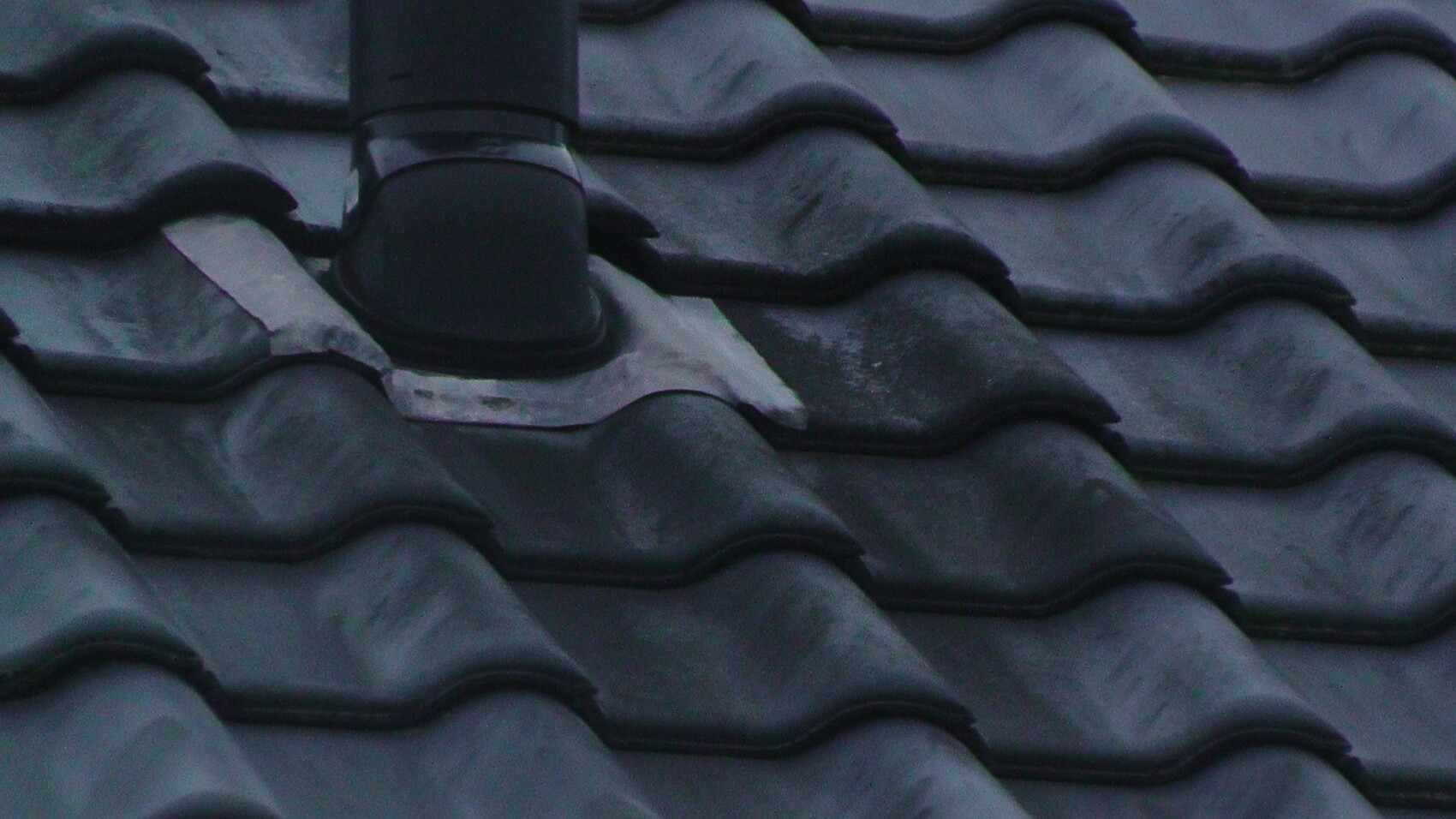 Close-up of a dark, wavy roof tile surface with pipe penetration and metal flashing