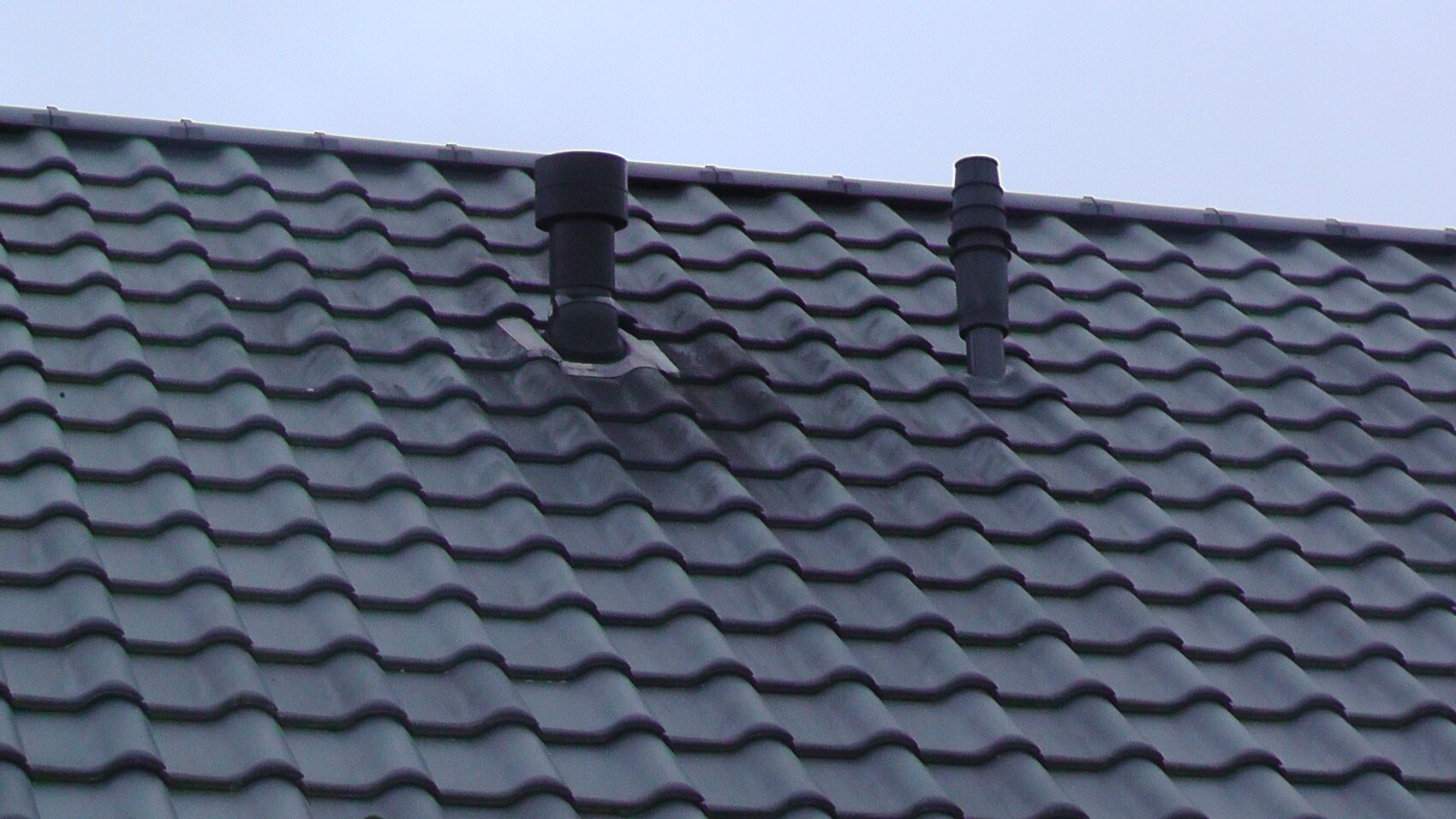 Roof with gray wavy tiles and two black chimney pipes against blue sky.