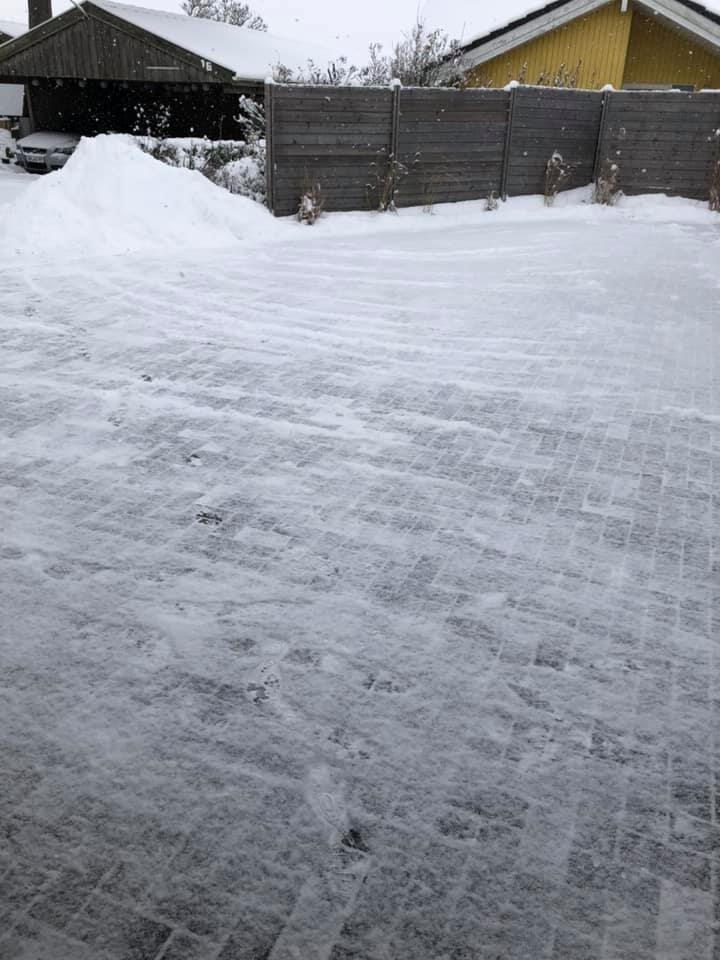 Snow-covered courtyard with wooden fence, garage on the left, yellow house on the right, light snowfall.
