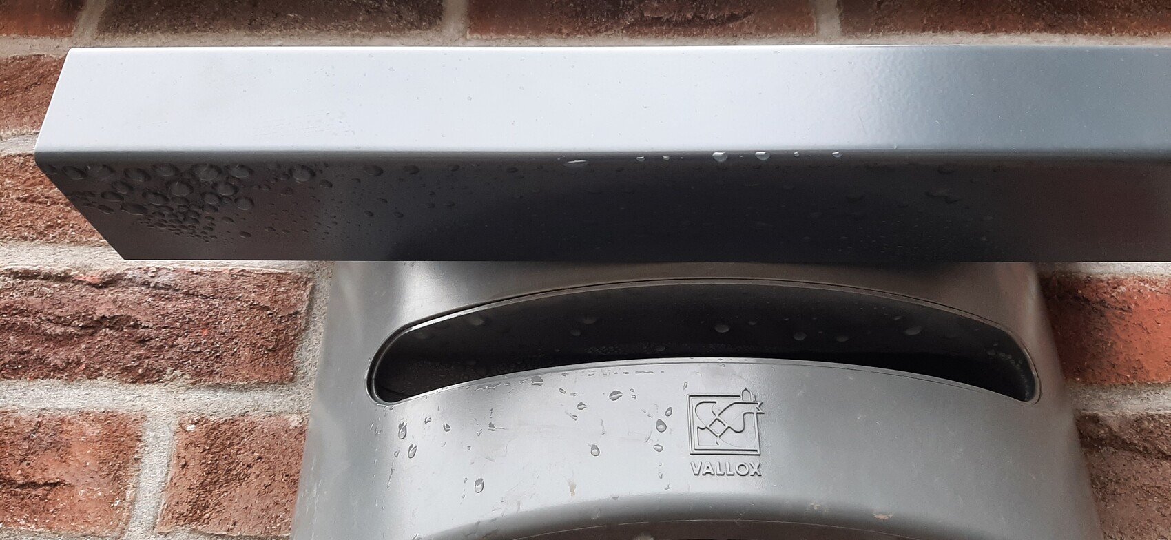 Gray metal casing on red brick wall, raindrops on the surface, VALLOX logo visible.