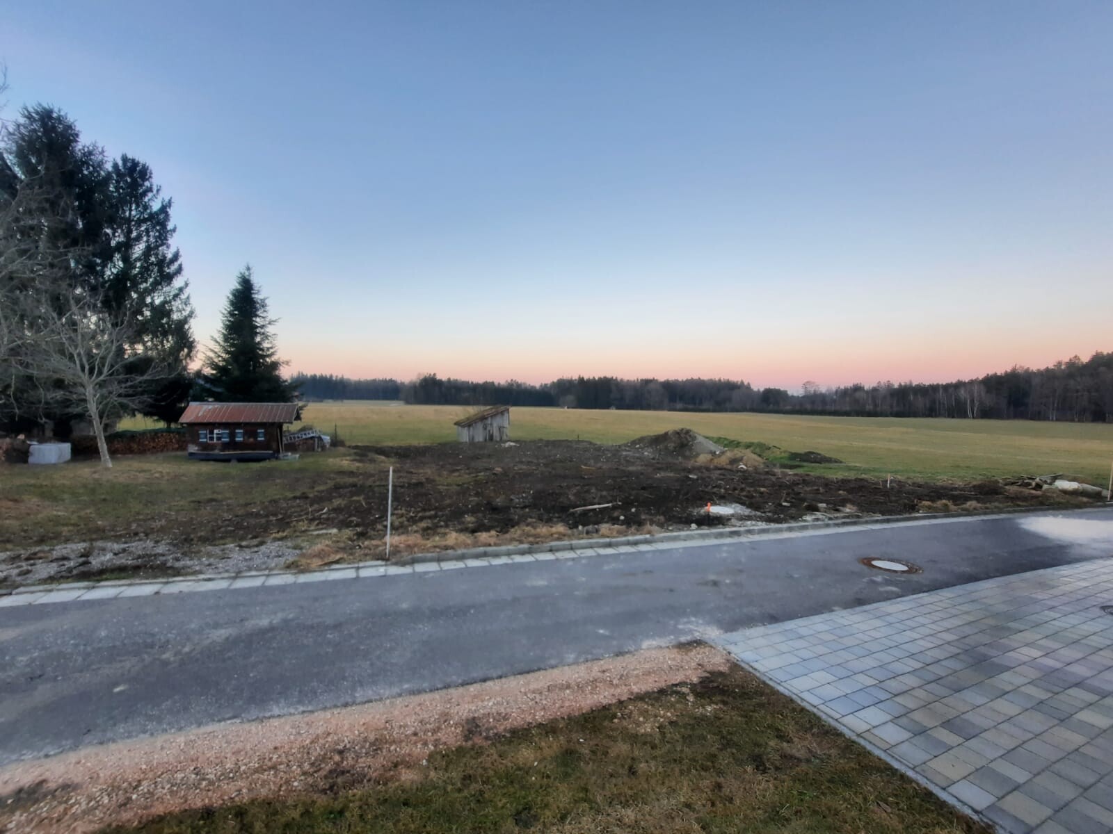 Sonnenuntergang über freiem Feld; linke Bäume, Holzhütte mit rotem Dach, Straße im Vordergrund