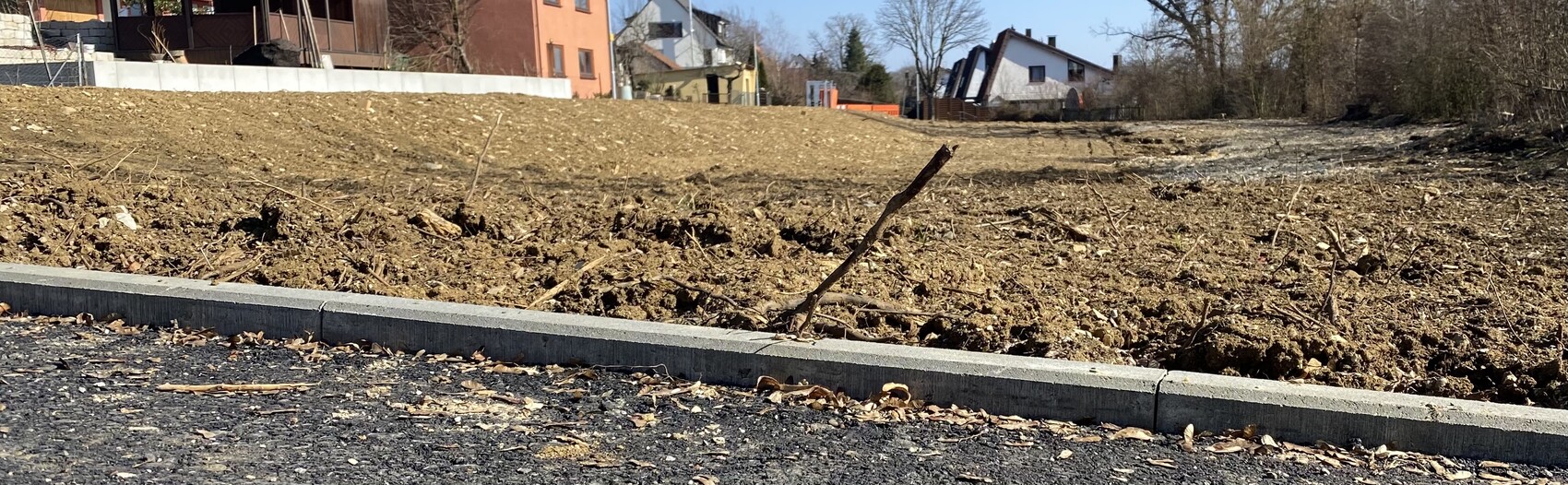Leeres Baugrundstück mit aufgeschütteter Erde, Asphaltstraße und Häuser im Hintergrund.