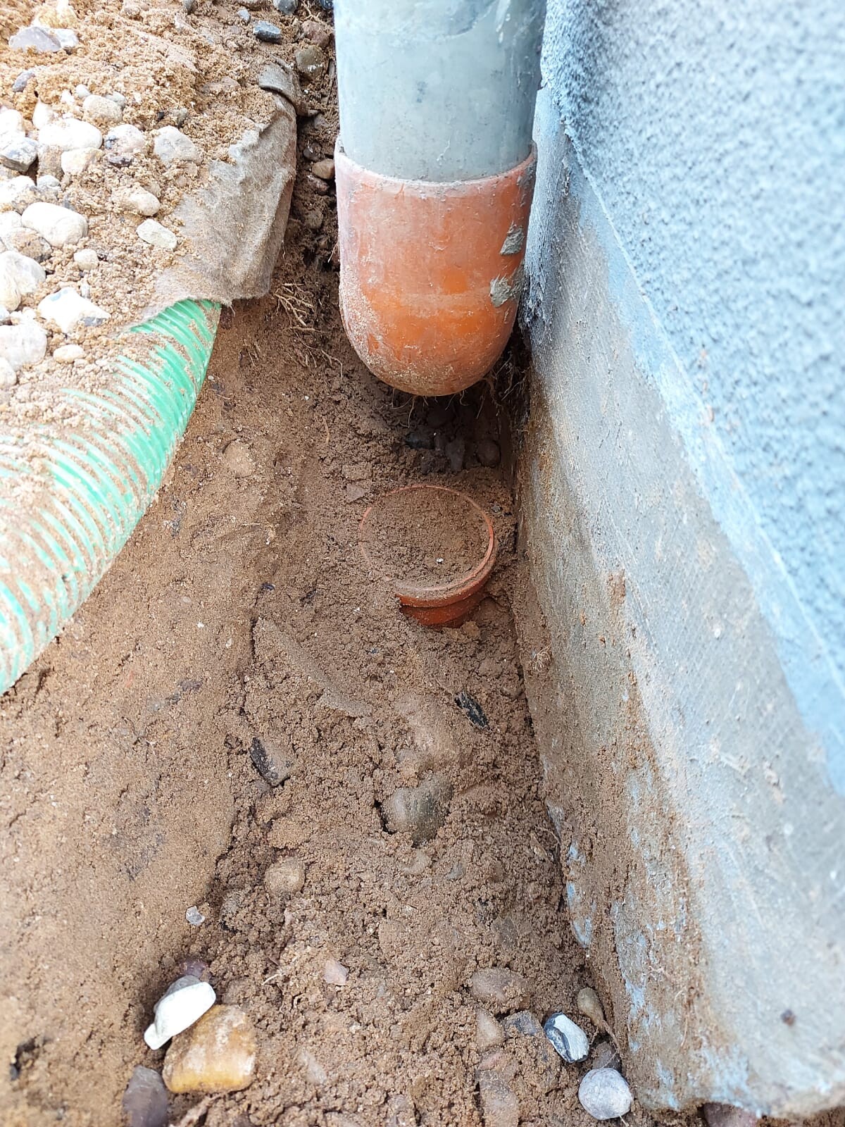 Drain pipe under the wall; orange connection, next to a green hose in sandy soil.