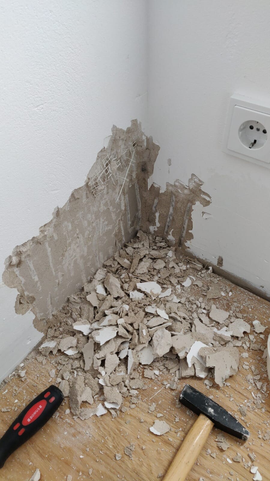 Broken plaster on the wall, rubble on the floor; hammer with a wooden handle next to an outlet.