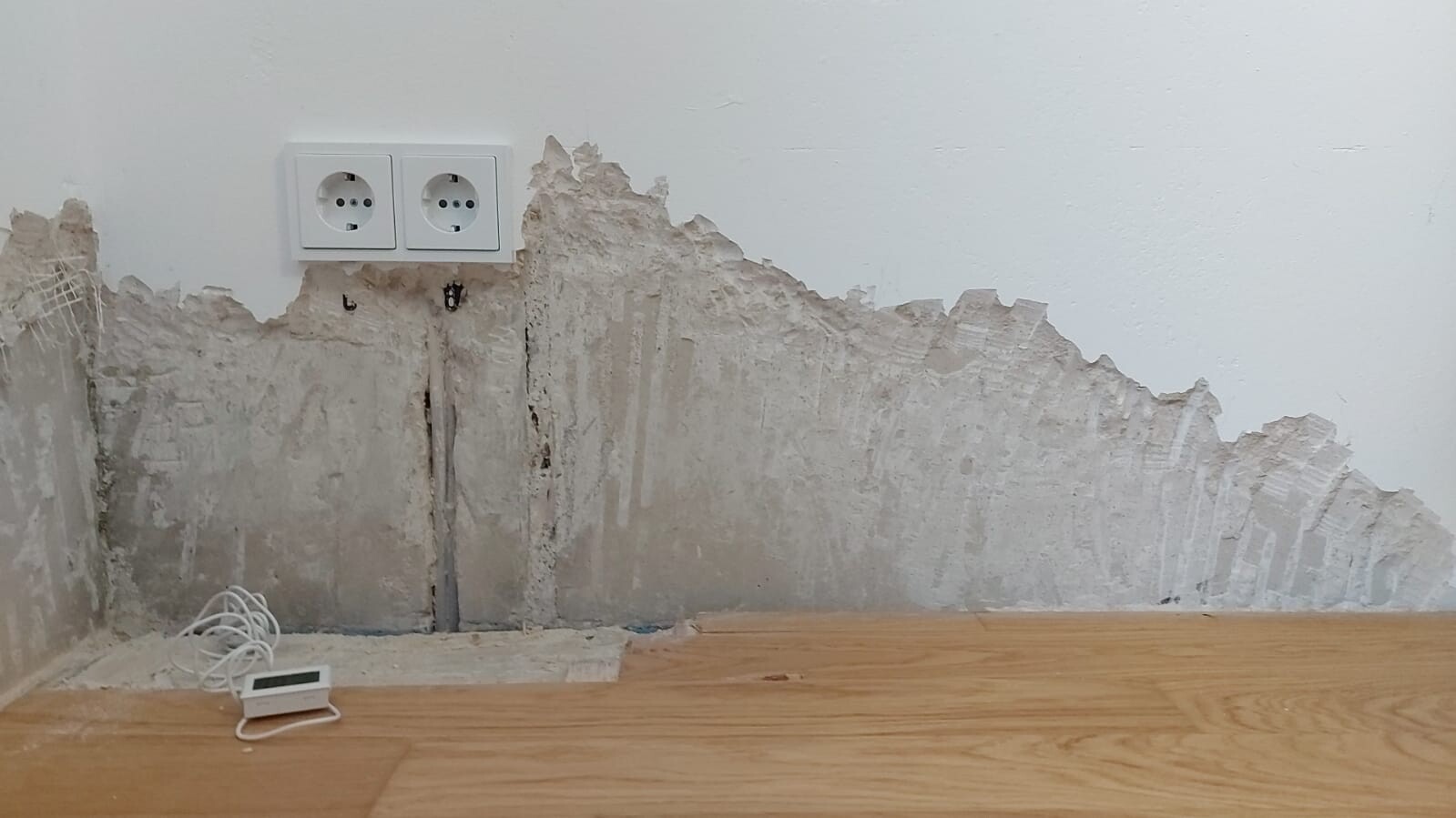 Damaged wall with exposed plaster, two outlets, loose cables and thermostat on wooden floor.