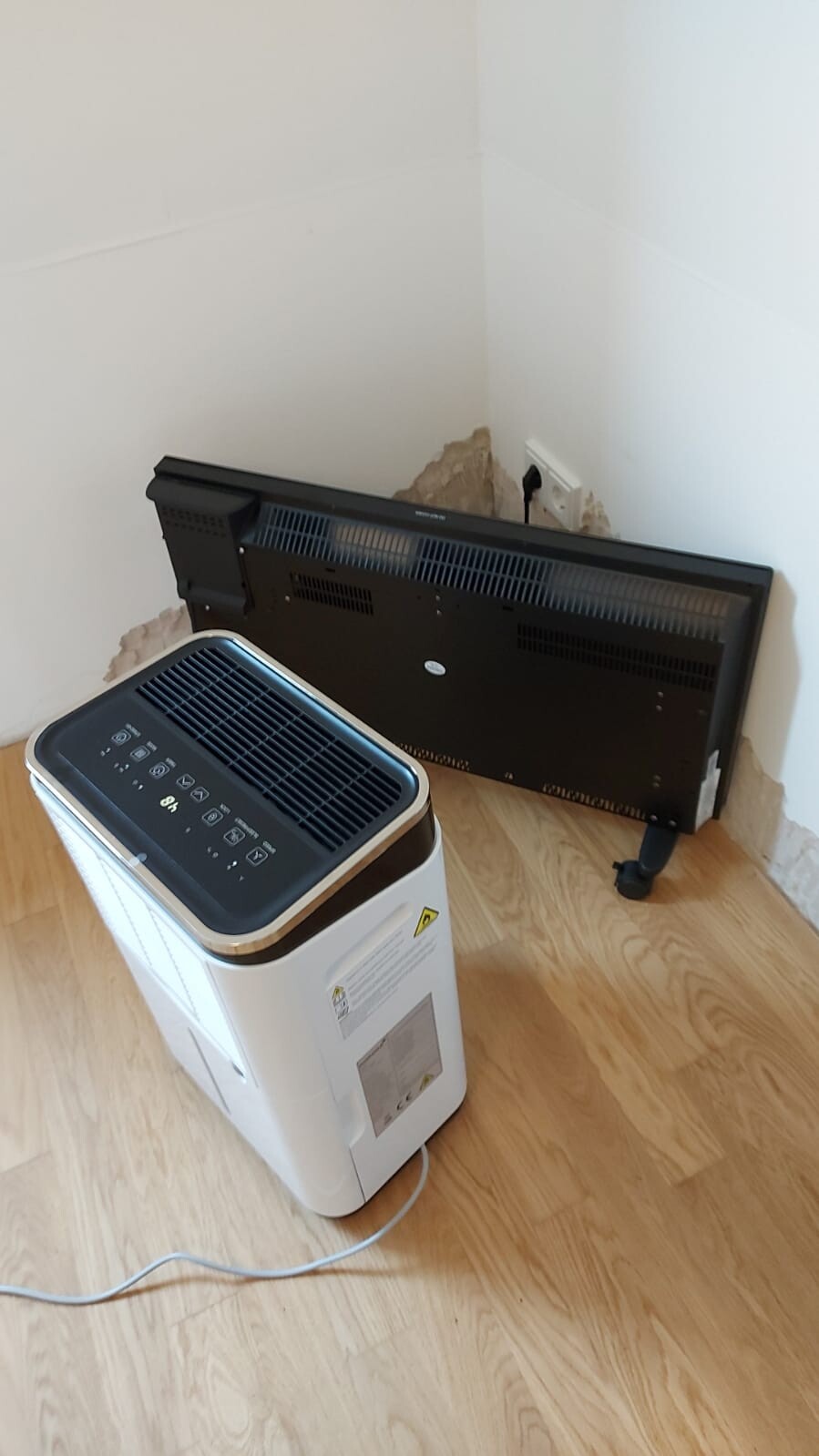 White dehumidifier on wooden floor; black fan heater on wall with plaster damage, visible cables.
