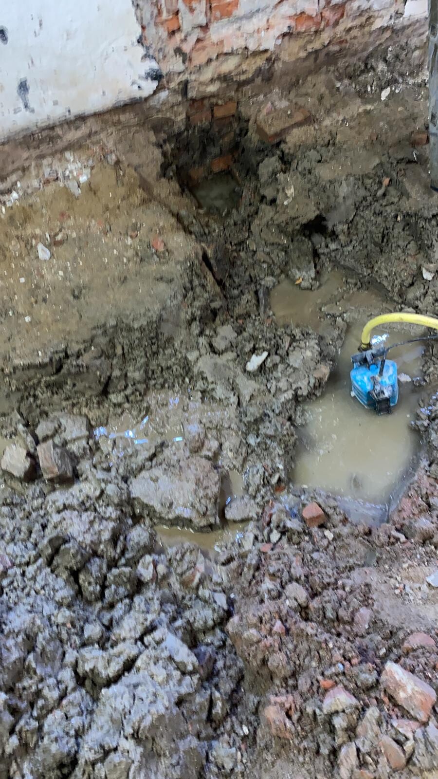 Construction pit with muddy ground, exposed brick wall and blue sump pump with yellow hose.