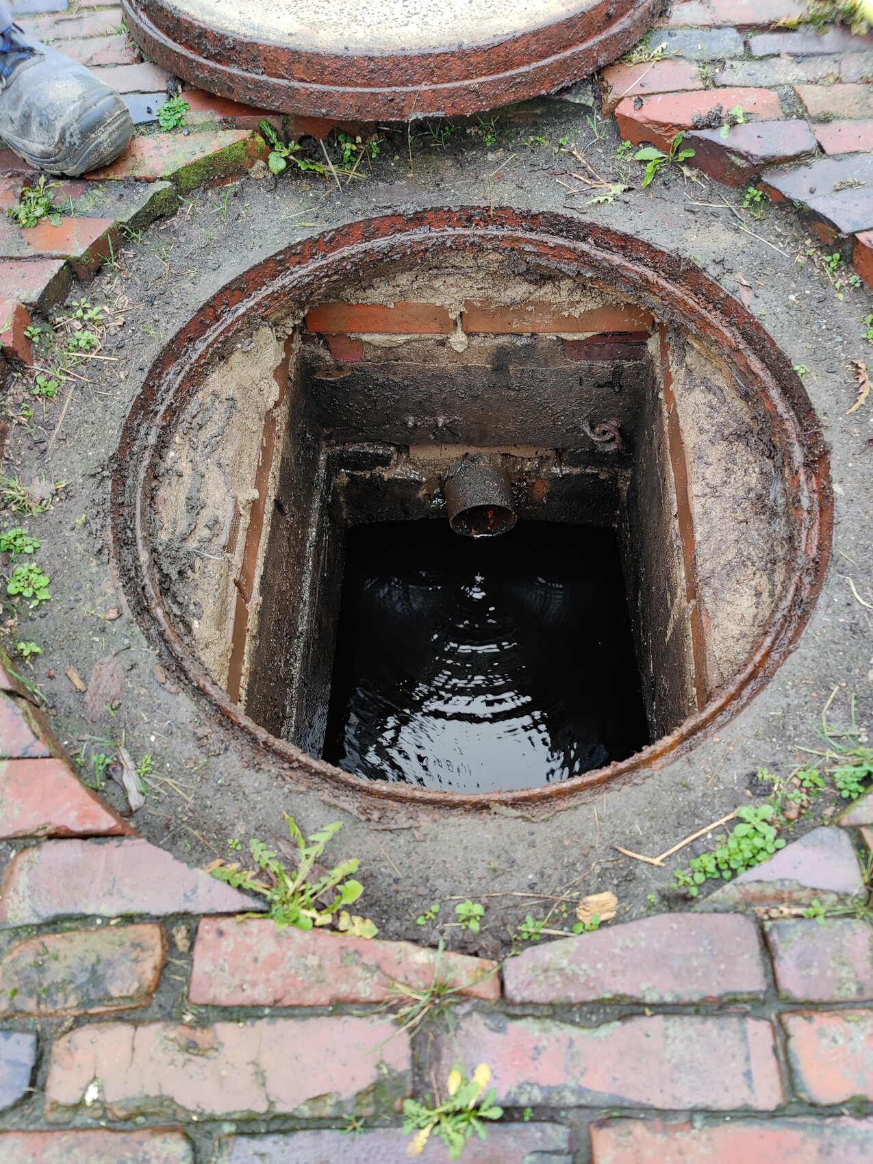 Open manhole shaft with dark water, rusty edge, surrounded by bricks and paving stones.