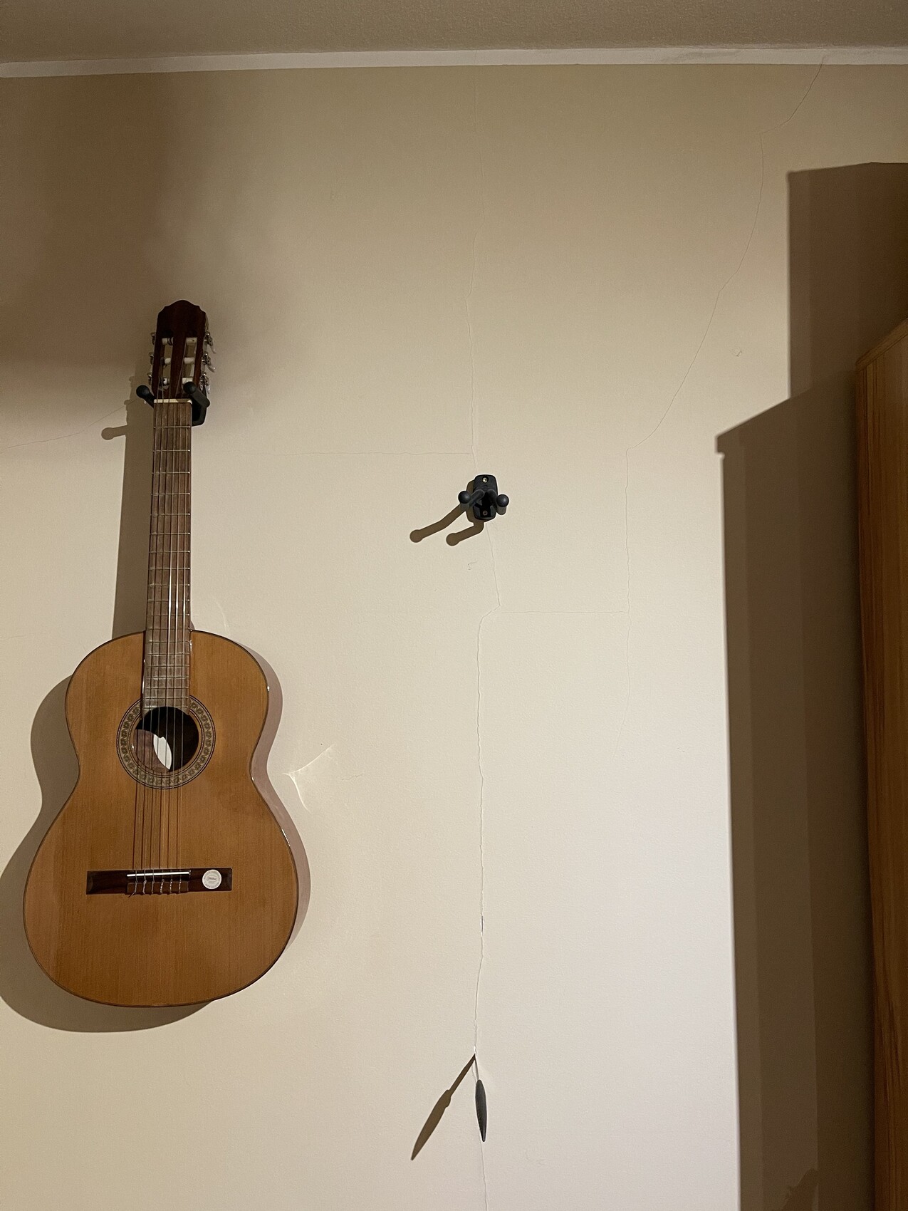 Acoustic guitar hanging on cream-colored wall with cracks; right cabinet visible.
