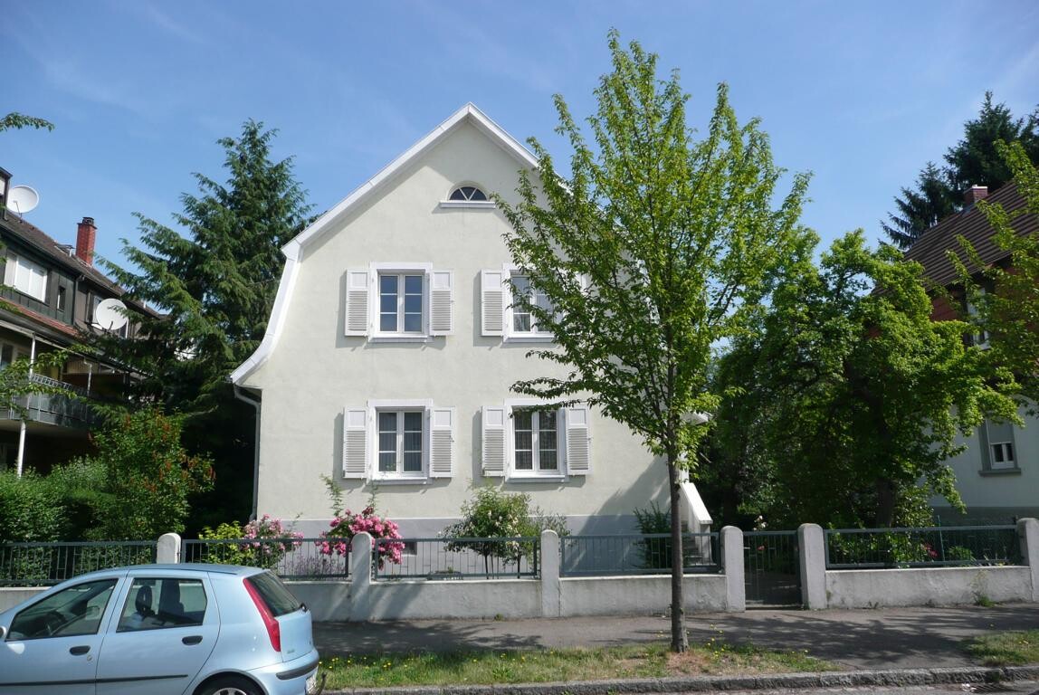 Exterior view of a light-colored house with white shutters, tree, and fence