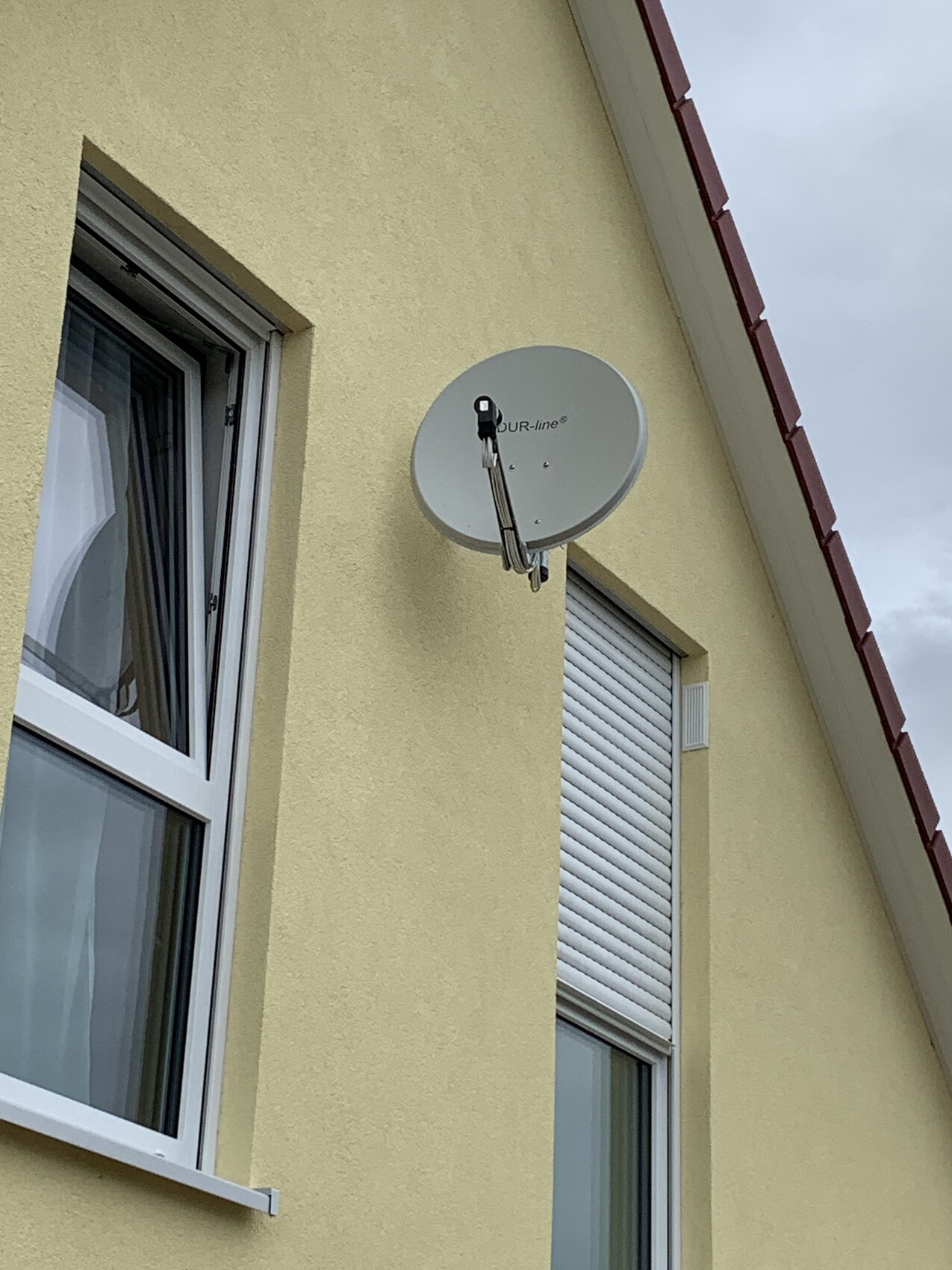 Satellite dish mounted on a yellow exterior wall above a window with roller shutter.