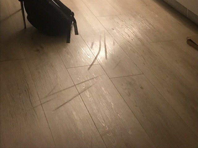 Holzboden mit mehreren Kratzlinien; eine schwarze Tasche liegt links oben.