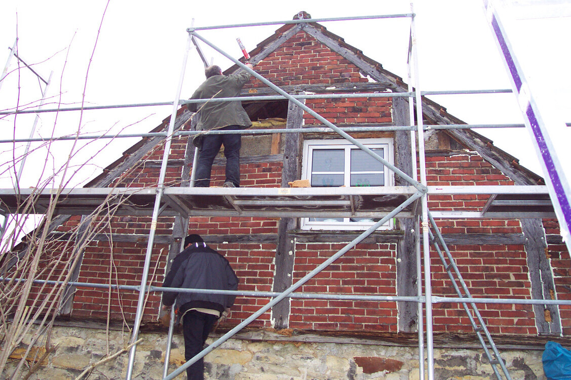Two construction workers standing on scaffolding in front of the red brick house roof; windows visible.