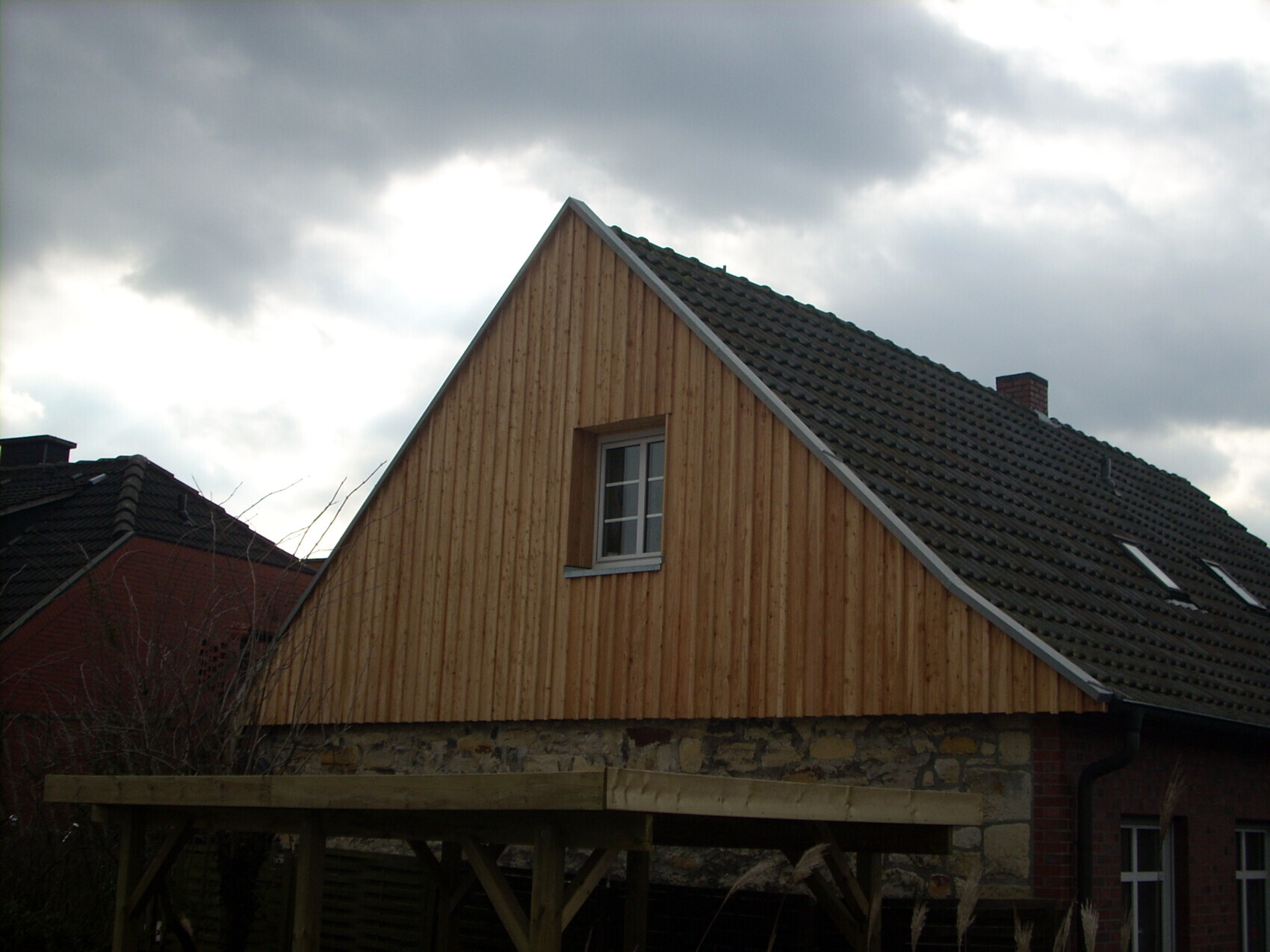 House with steep wooden gable, dark tiled roof and stone foundation against cloudy sky.