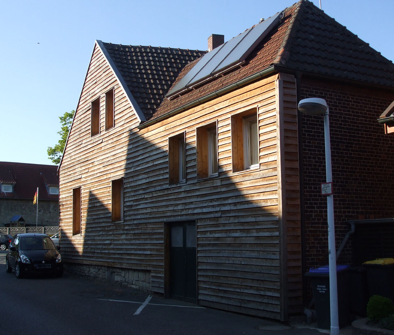 Wooden house with solar panels on the roof; street scene with street lamp and parked car