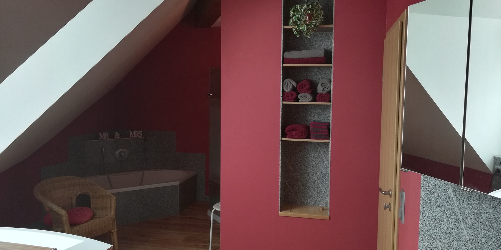 Wellness bathroom in the attic with red wall, corner bathtub, shelf with towels and wicker chair.