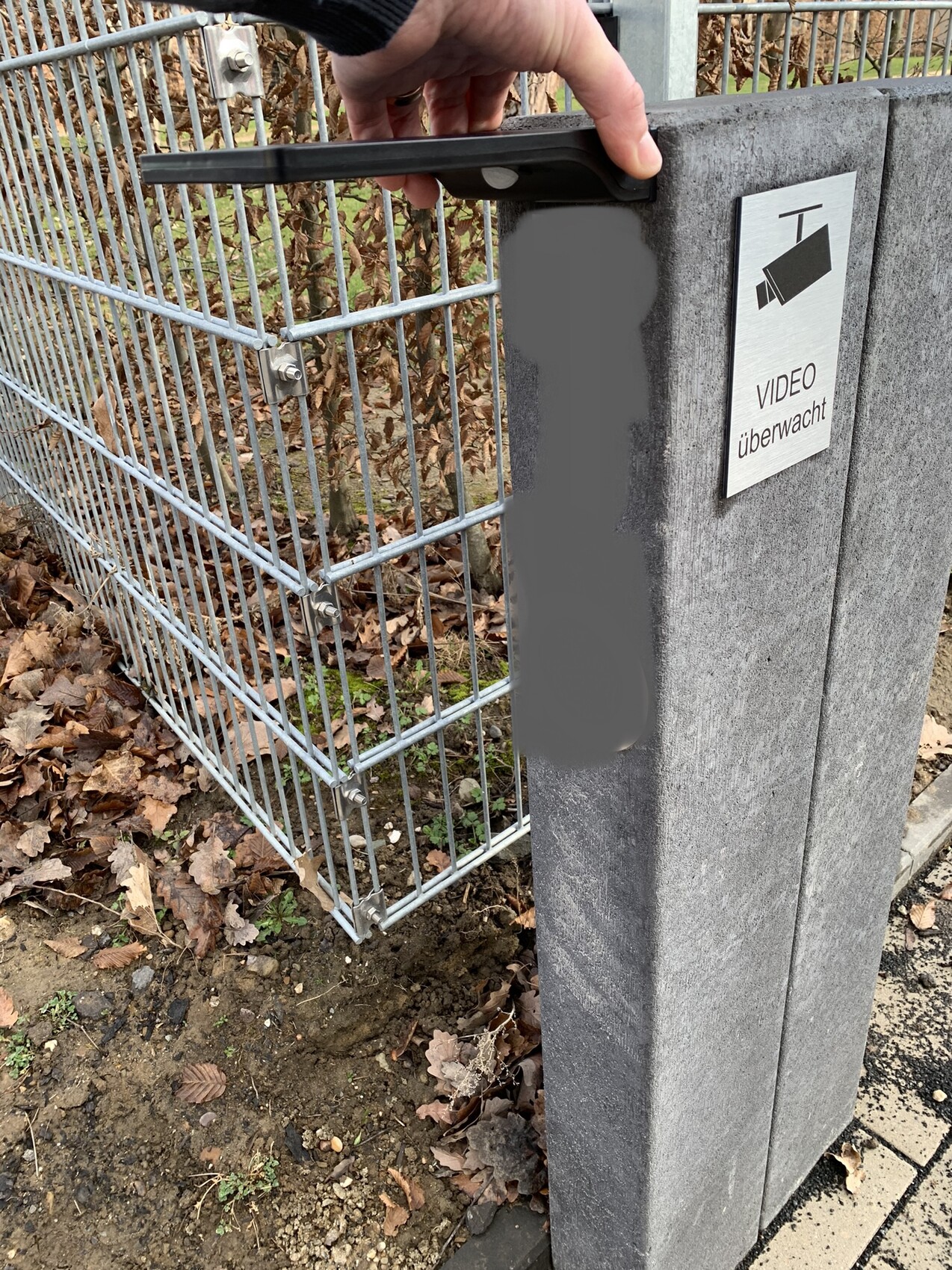 A hand opens metal gate on concrete post; visible sign saying 'Video monitored'.