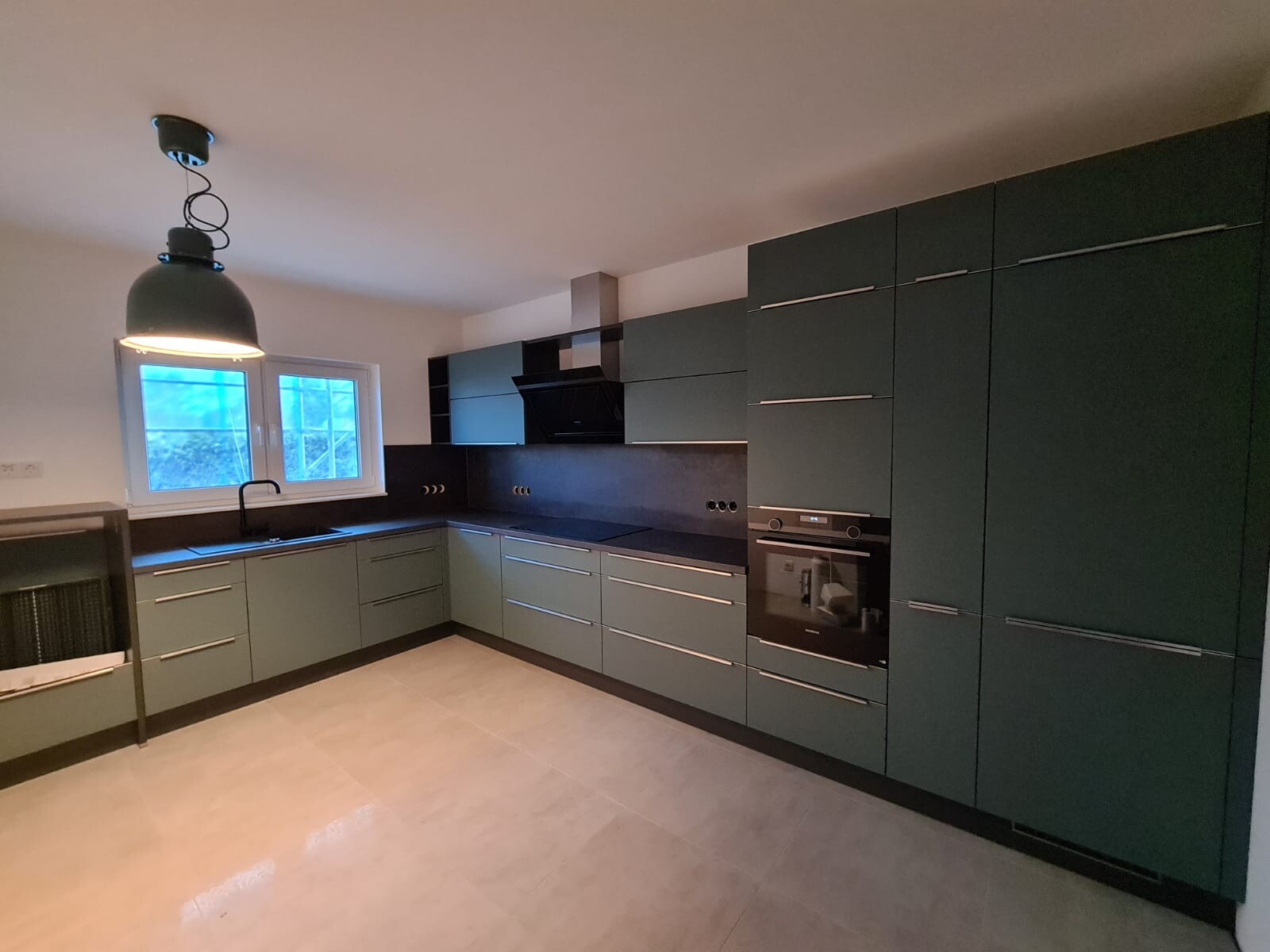 Modern kitchen with green fronts, black countertop, oven, extractor hood, and sink by the window.