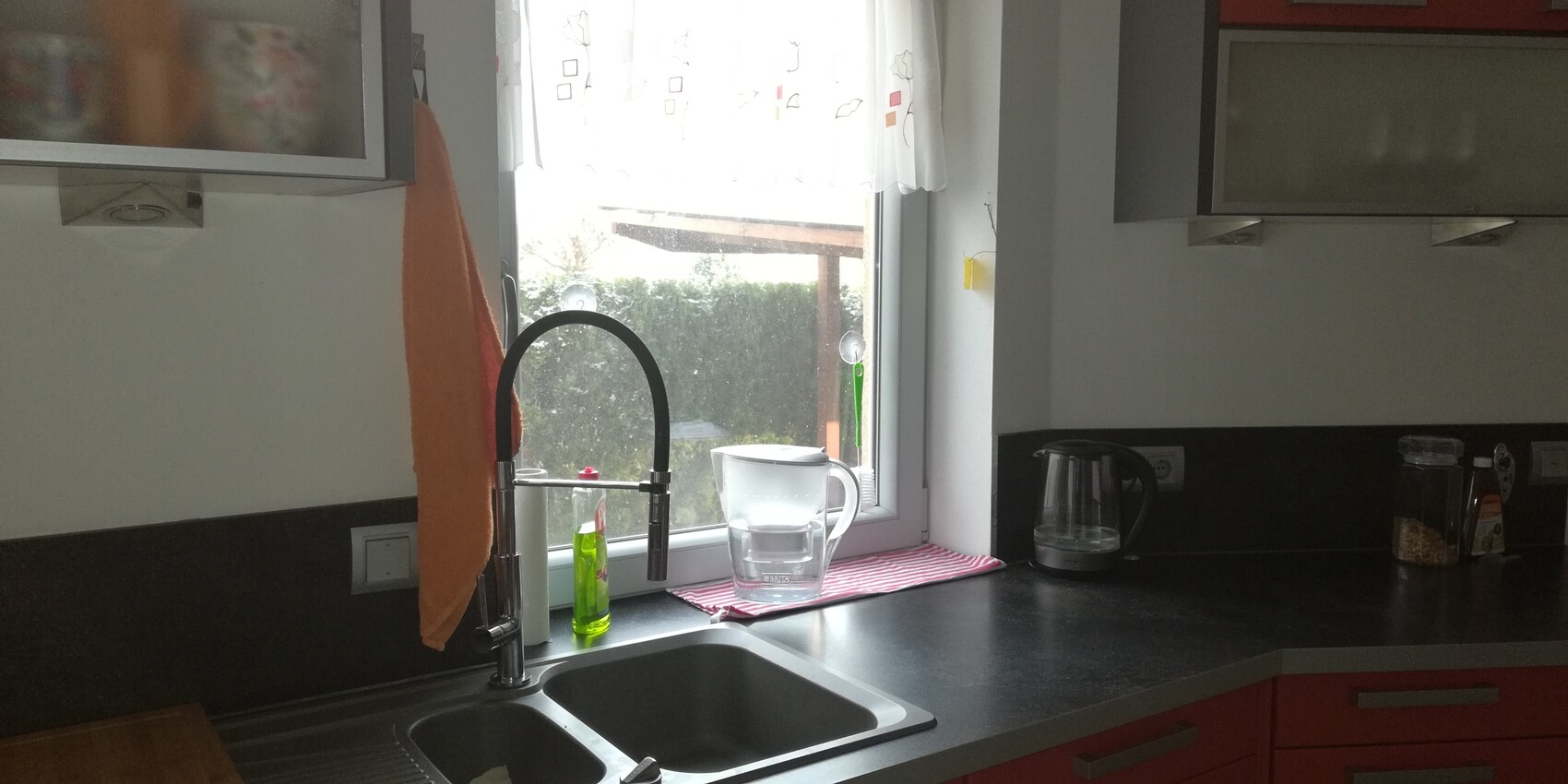 Kitchen with sink by the window: high-arc faucet, green dish soap bottle, glass carafe, orange towel.