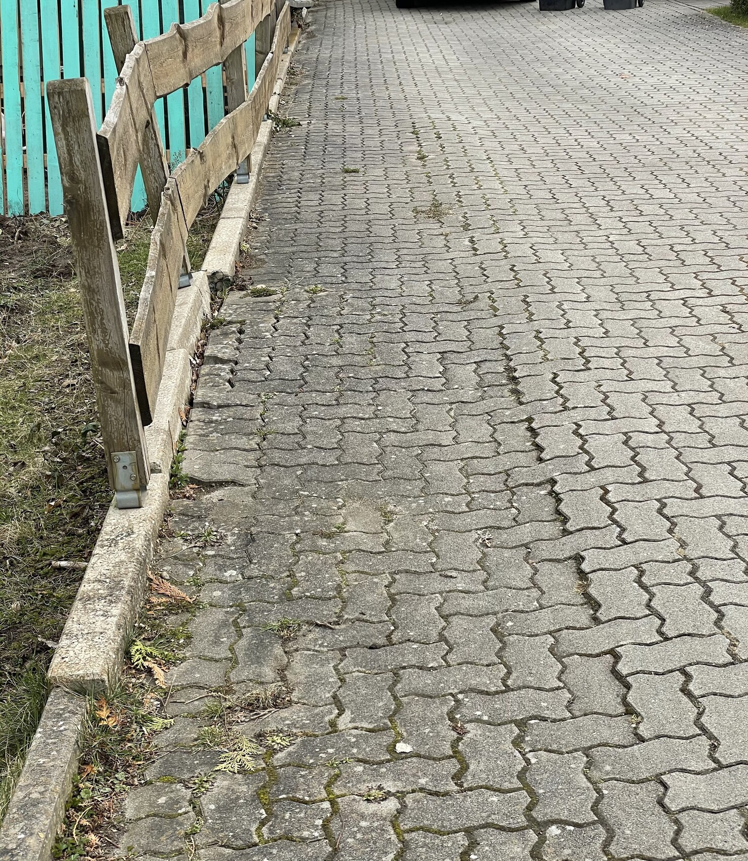 Paved walkway made of concrete pavers with wooden slat fence on the left; turquoise fence behind.
