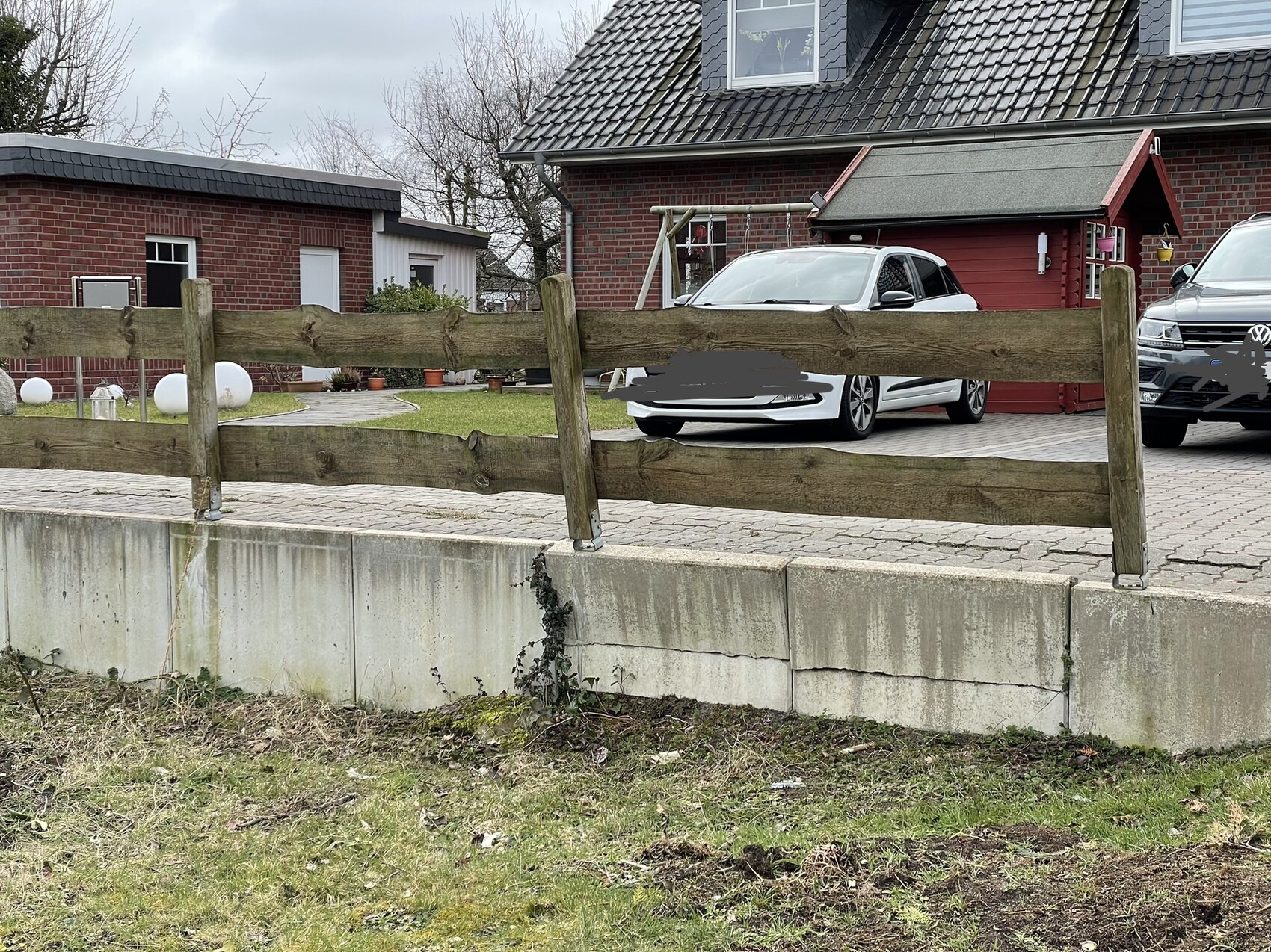 Wooden slat fence in front of a concrete wall, with a white car and grey SUV in front of a red house behind.