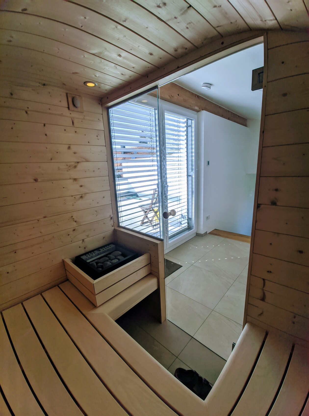 Wooden sauna with curved benches, sauna stove and glass door to the outside area with blinds.