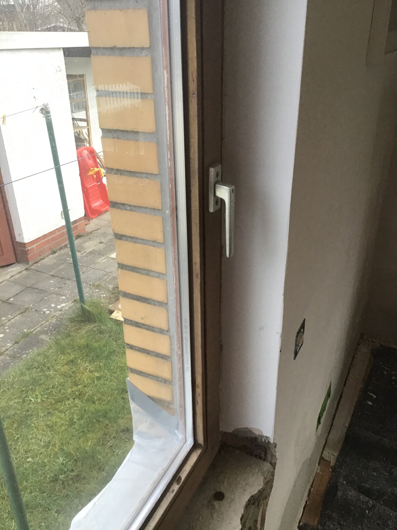 Blick durch Fenster: außen gelbe Ziegelwand, roter Plastikstuhl, Wäscheleine; innen Wandloch.
