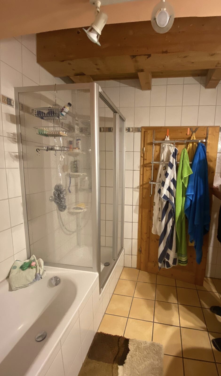 Badezimmer mit Glasdusche, Badewanne links, Holzbalkendecke und bunte Handtücher an der Tür.