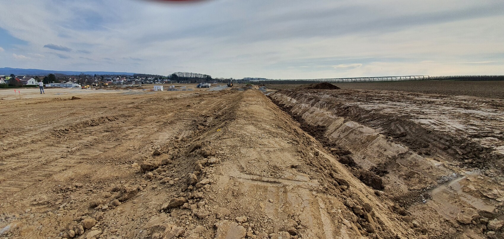 Large construction site with trenches, earthworks and construction vehicles; village and fields in the background.