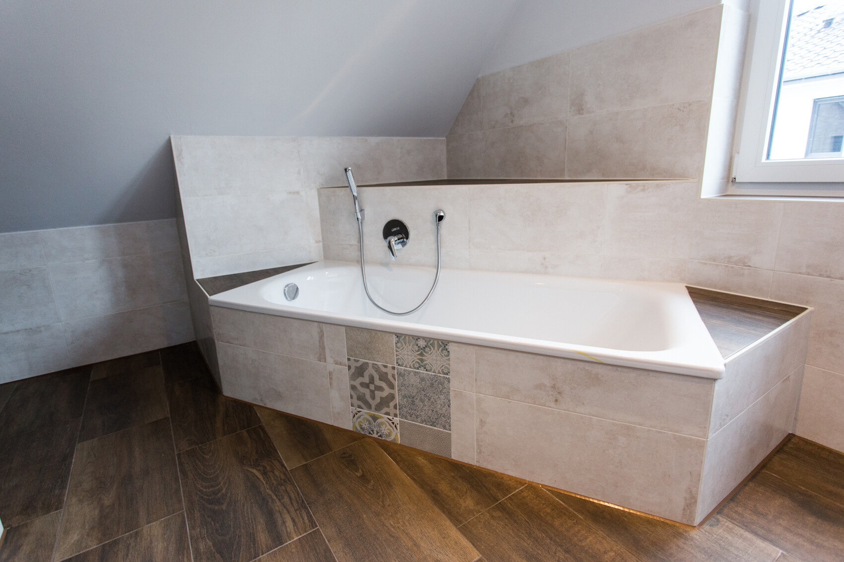 Modern bathtub with beige tiles, handheld shower, by the window, wooden floor.