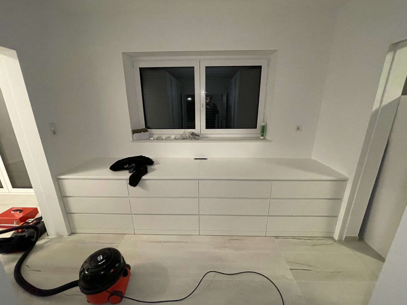 White, modern dresser under window; black clothing item lying on it, vacuum cleaner on the floor.