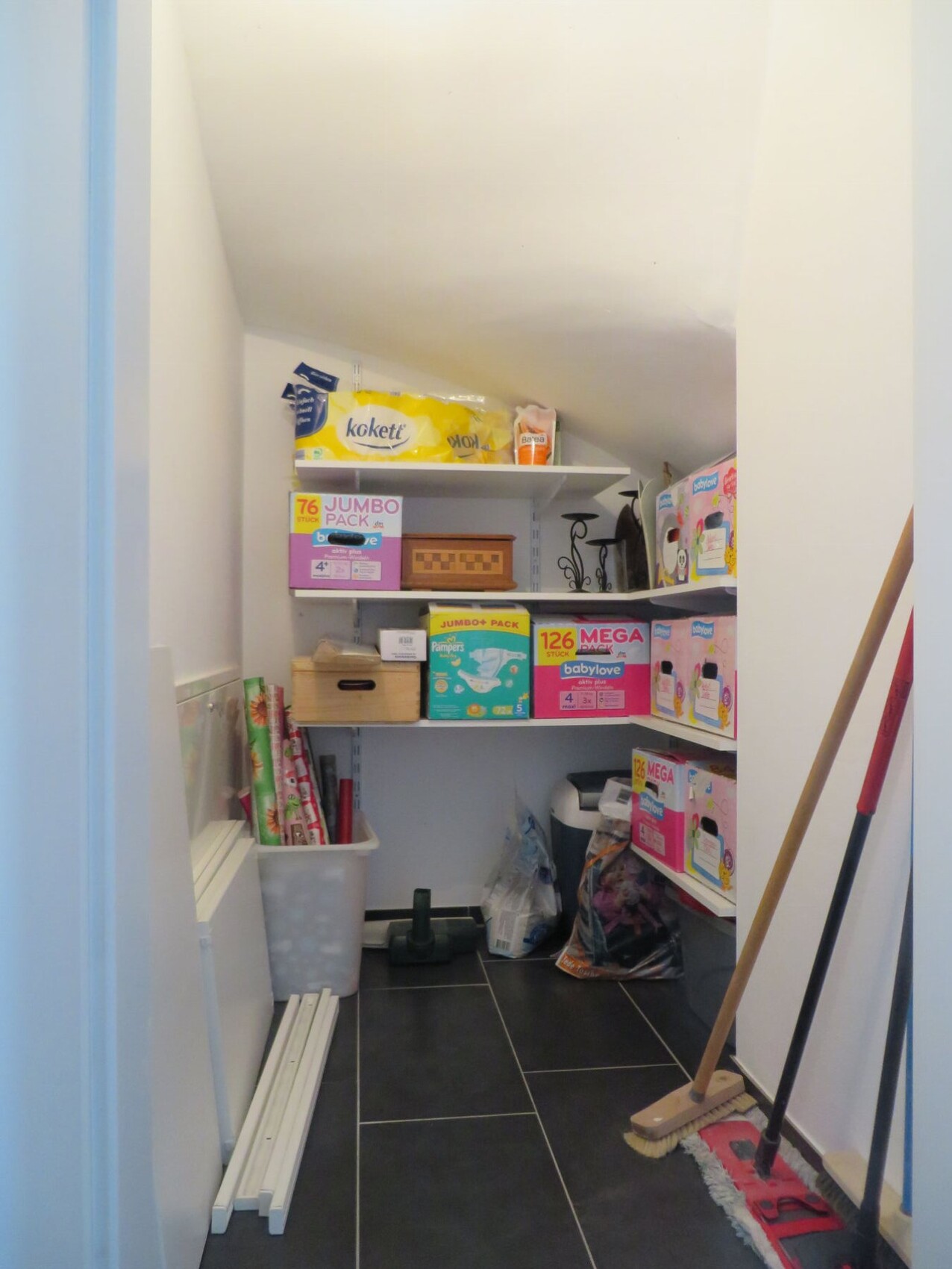 Small storage room with shelves full of diapers, cleaning products, brooms, and mop.