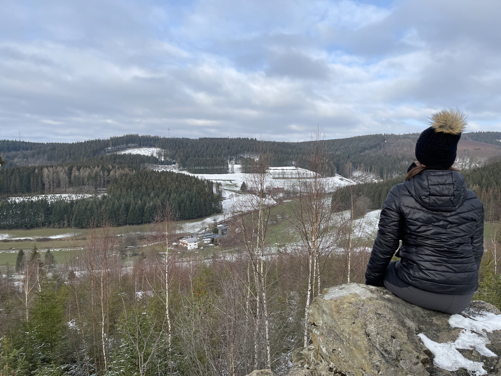 Person sitting on rocks looking over snow-covered forest and meadow landscape with houses.