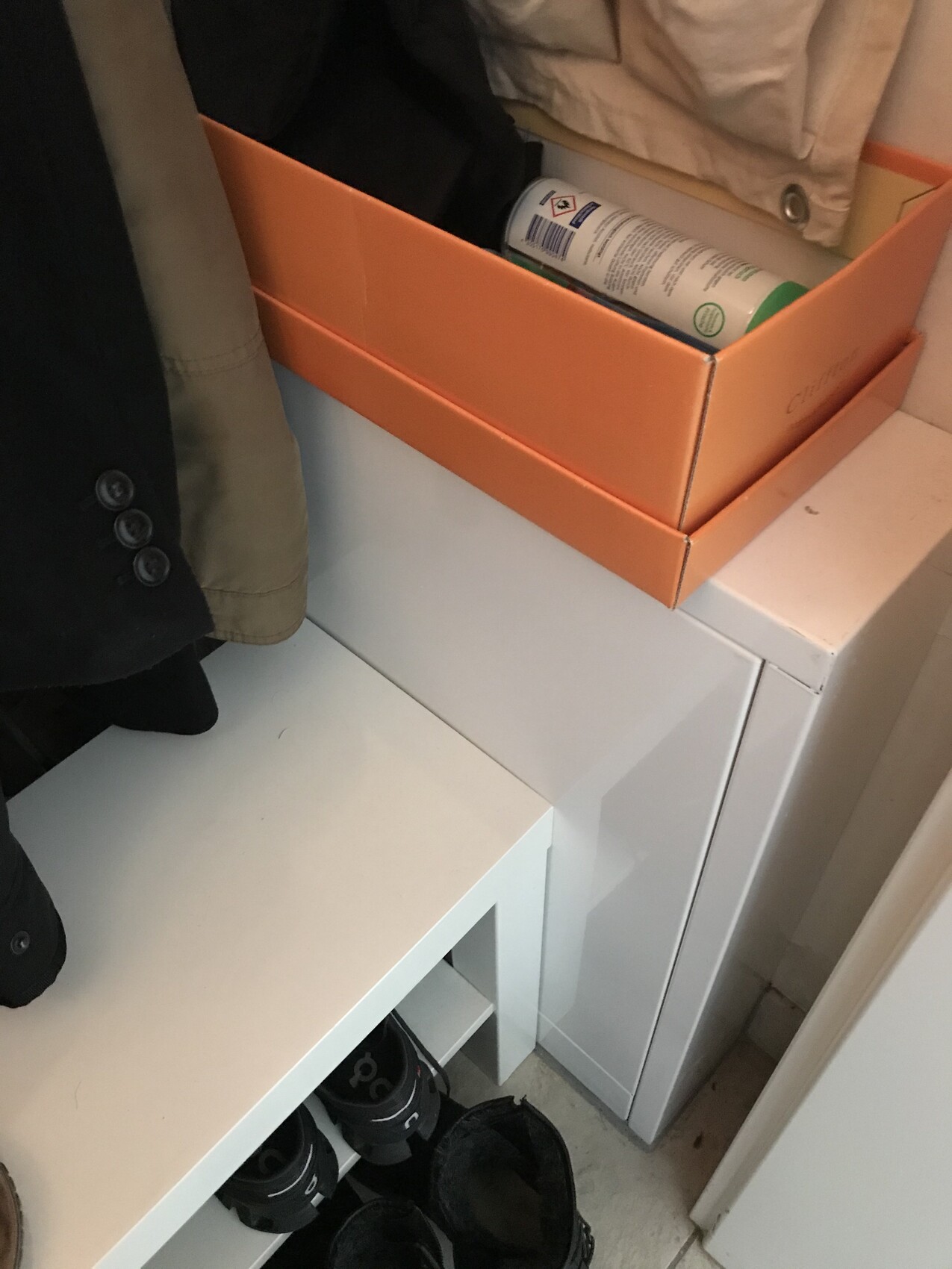 Orange box with spray can on a washing machine; coat hanging on the left, shoes below.