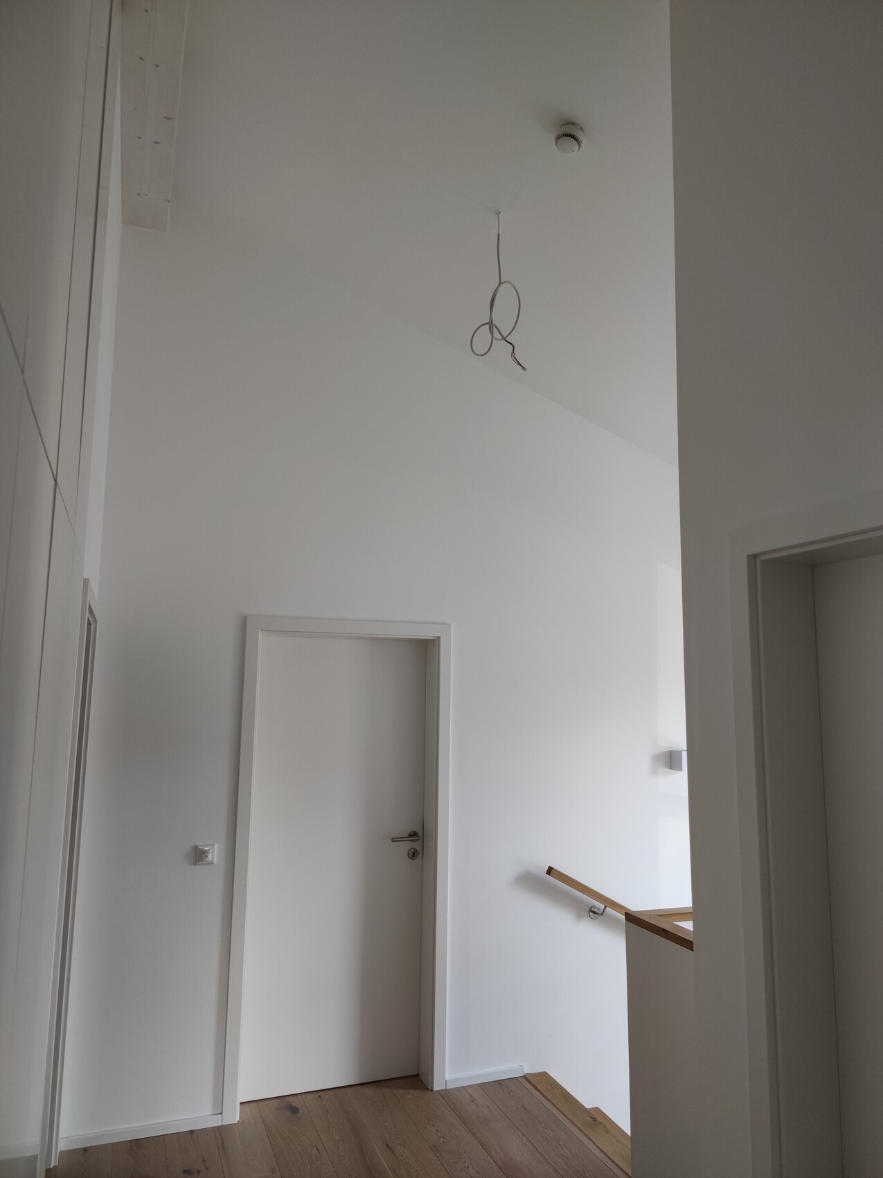 Bright hallway/stairwell with white walls, open cable from the ceiling, door on the left, wooden floor.