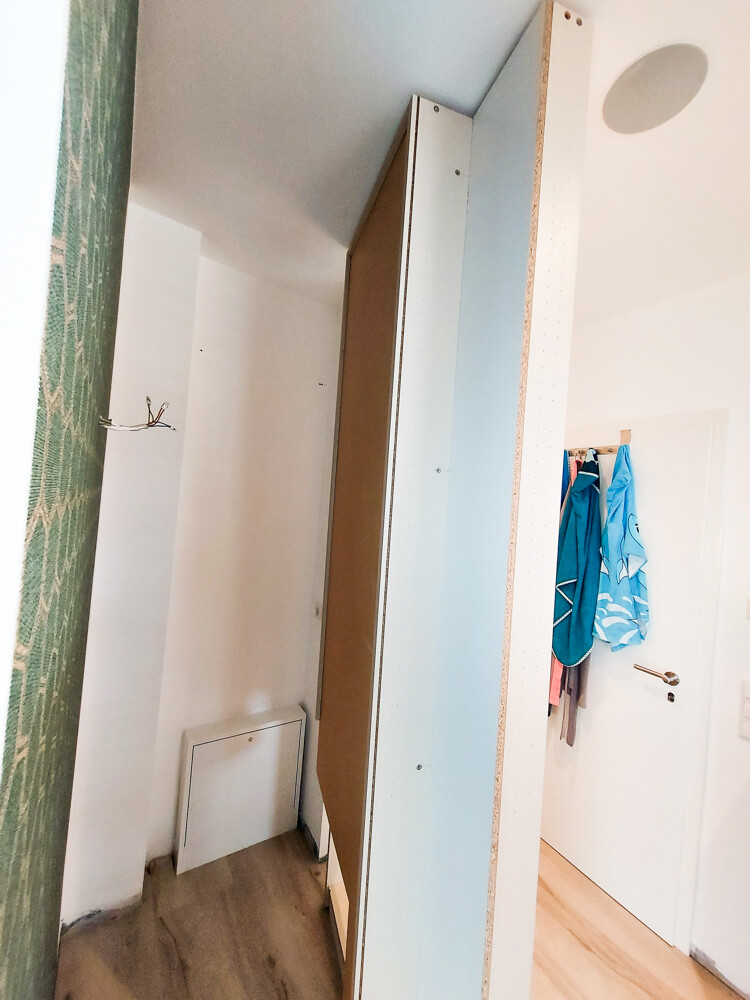 Freestanding, narrow furniture piece made of wooden panels; white walls, wooden floor, blue jackets.