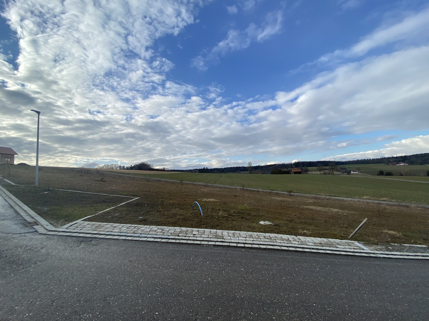 Straßenrand mit Laterne, Gehwegplatten, freies Baugrundstück, Felder und Himmel mit Wolken.