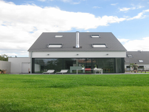 Modern house with large glass front, skylights, patio furniture, and green lawn.