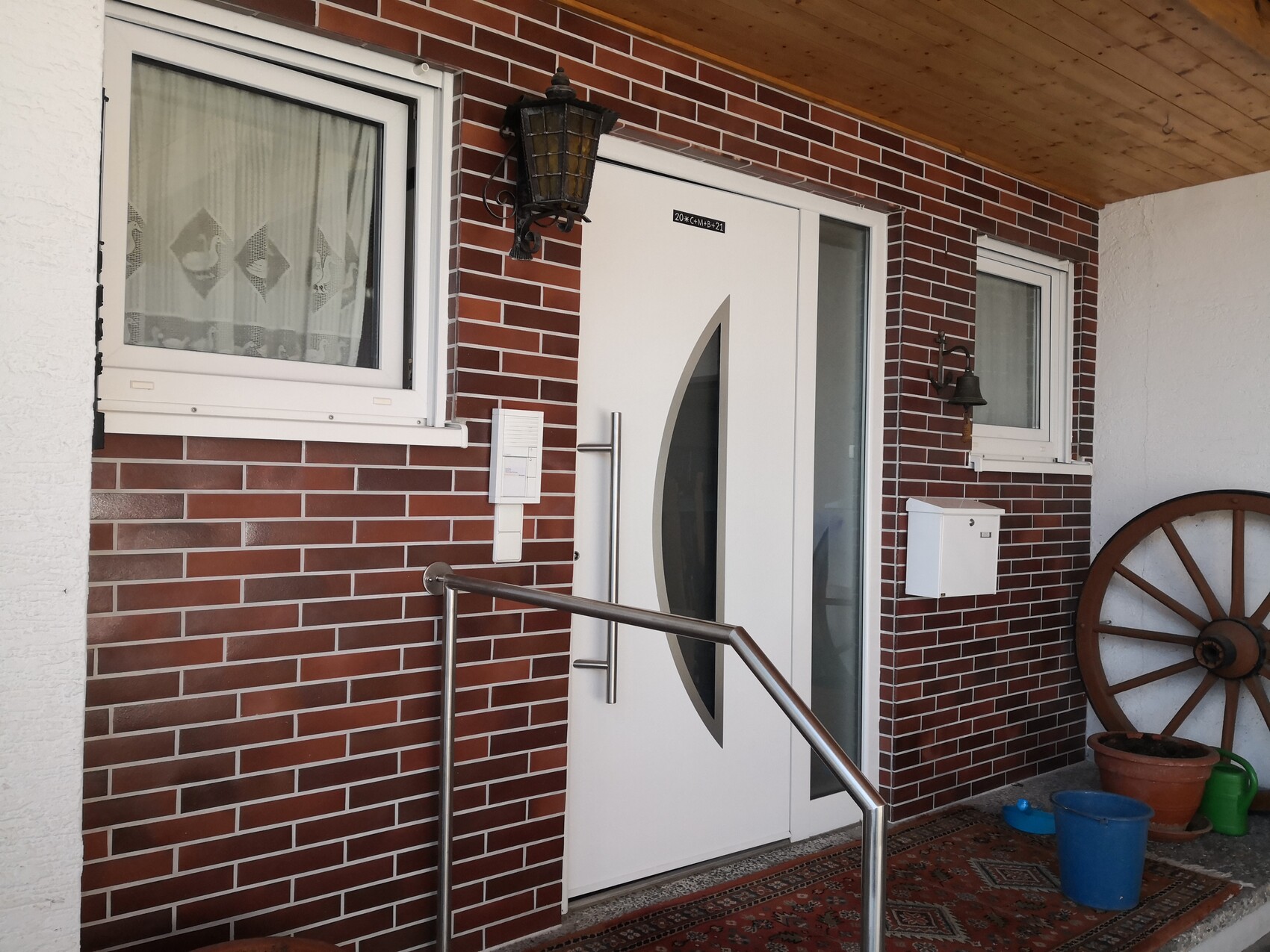 White door with oval glass window at red brick entrance; left window, lantern, wagon wheel.