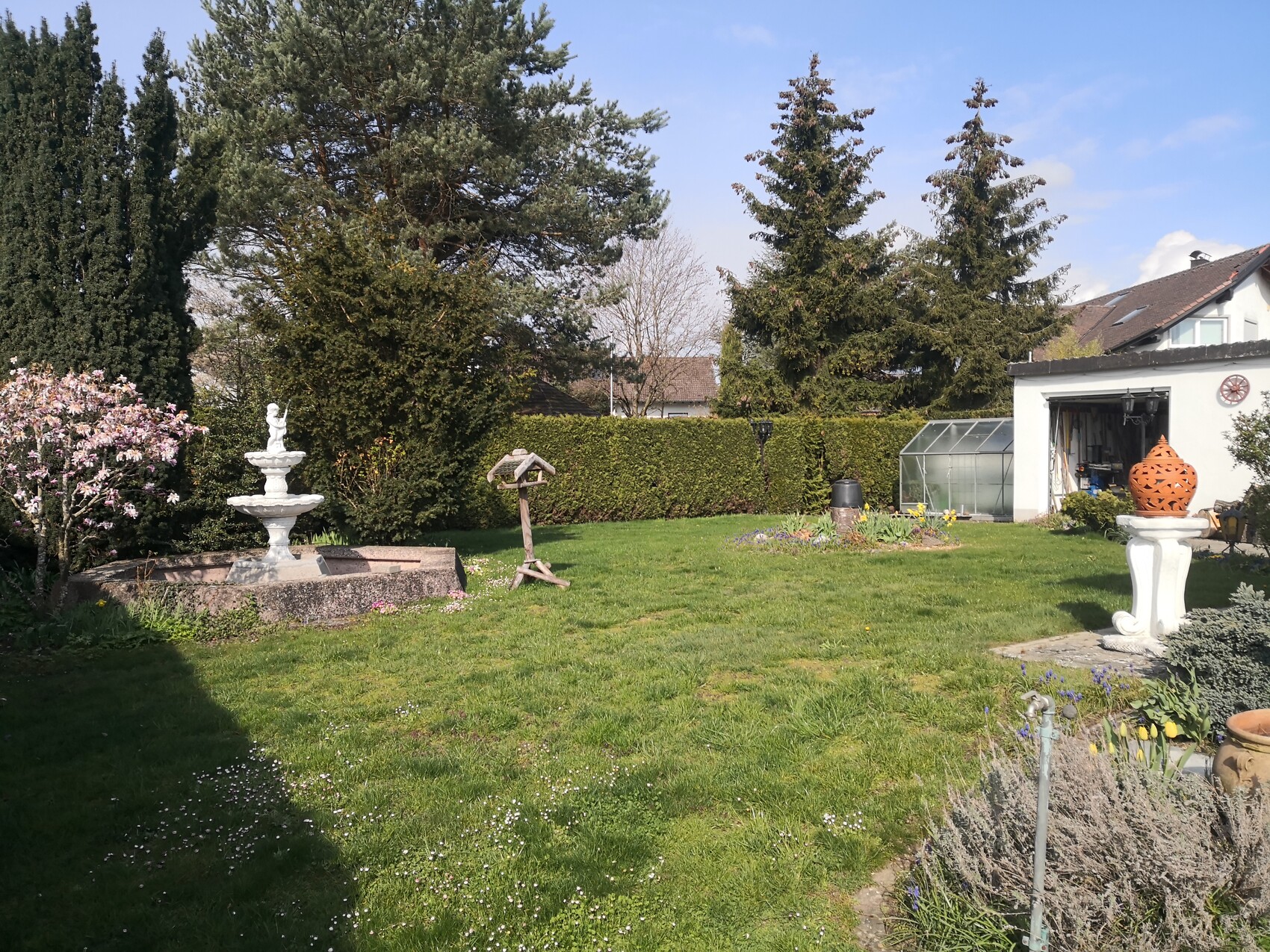 Garden with fountain statue, green lawn, greenhouse and orange decorative ball on pedestal.